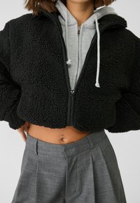 Musta, lyhyt takki, joka on valmistettu teksturoidusta, pörröisestä materiaalista ja jossa on vetoketju edessä; päällä harmaa hupullinen collegepaita ja korkeavyötäröiset harmaat housut.