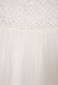 Robe blanche avec un corsage en dentelle florale et une jupe en chiffon plissée douce. Le design présente une superposition texturée en dentelle avec un tissu lisse en dessous.