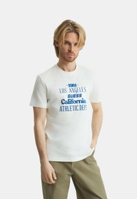 T-shirt in cotone bianco con stampa grafica blu e testo: "1981 LOS ANGELES GUESS California. ATHLETIC DEPT." Maniche corte, colletto tondo.