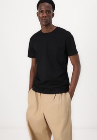 Black short-sleeve t-shirt made of cotton, featuring a small logo on the chest, paired with beige loose-fitting pants. Plain background.