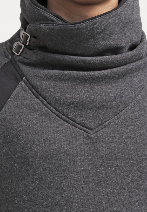 Gros plan d'une personne portant un pull à col montant gris foncé avec deux boucles de sangle noires sur le col.