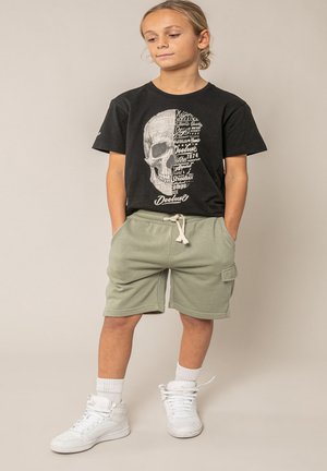 Kind staat met handen in zakken en draagt een zwart T-shirt met schedelprint, lichtgroene korte broek met trekkoord, witte sokken en witte hoge sneakers.