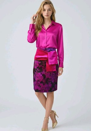 Mujer con cabello rubio largo, vestida con una blusa de satén rosa brillante, falda floral morada y rosa, y tacones altos dorados, de pie con una mano cerca de su cara.
