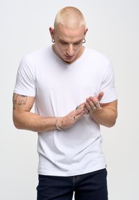 Bílý v-neck tričko, měkká tkanina, krátké rukávy, nošeno s tmavými džíny. Ruce zdobené několika prsteny, zkoumá jednu ruku s druhou.