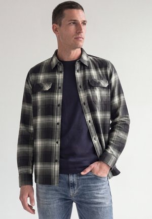 Mann trägt ein schwarz-grau kariertes Hemd mit Knöpfen über einem dunkelblauen T-Shirt und hellblauen Jeans, steht mit einer Hand in der Tasche.