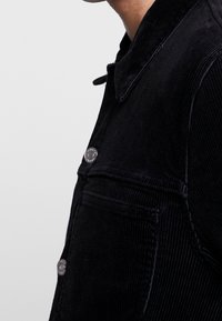 Svart manchesterjacka med en strukturerad krage, synliga sömmar och metallknappar med tryckknappsfäste. Har sidofickor och bröstfickor.