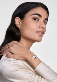 Montre-bracelet en mailles dorées avec un cadran blanc, accompagnée d'un bracelet délicat orné de petites pierres claires, portée sur un bras à la peau claire.