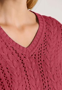 Nahaufnahme einer Person, die einen rosa gestrickten V-Ausschnitt-Pullover mit einem Zopfmuster und Lochmuster trägt.