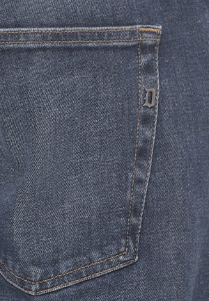 Bolsillo trasero de pantalón vaquero con costuras naranjas y una pequeña letra metálica "D" adherida a la costura del lado derecho del bolsillo.