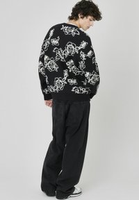 Pull noir oversize avec des motifs floraux blancs, tricot texturé, poignets côtelés, associé à un jean ample foncé et des baskets blanches.