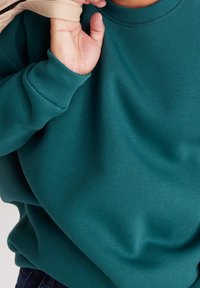 Bluza w kolorze morskim z prążkowanymi ściągaczami przy mankietach i dekolcie, o gładkiej teksturze. Ręka trzyma częściowo widoczny jasny materiał.