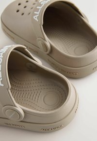 Sabots en caoutchouc beige avec une intérieur texturé et un logo sur la sangle. Ils présentent des trous ronds pour la ventilation et un design lisse et minimaliste.