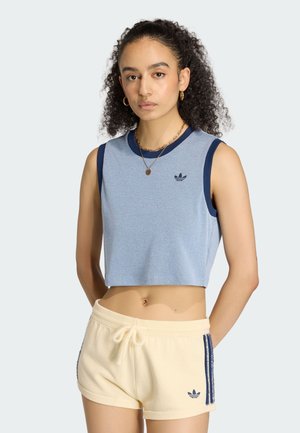 Młoda kobieta o kręconych włosach nosi jasnoniebieski bezrękawnik typu crop top i kremowe szorty z granatowymi paskami, stojąc na jednolitym tle.