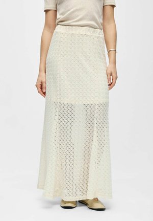 Femme portant une longue jupe crème en crochet par-dessus un jupon beige, associée à un top beige clair à manches courtes et des chaussures beige sans lacets.