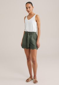 Green linen shorts with a scalloped, embroidered hem and self-tie belt, paired with a white sleeveless v-neck top. Smooth texture.