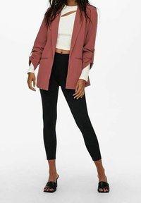 Kvinna i rostfärgad kavaj, vit kortärmad topp med utskärning, svarta leggings och svarta öppna klackar som står mot en vit bakgrund.