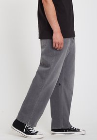 Pantalones vaqueros grises con un corte holgado, ligera desgaste, bolsillos laterales, combinados con zapatillas negras de corte bajo.