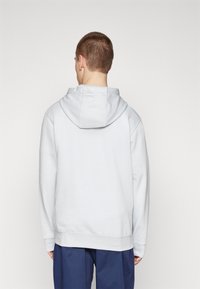Ljusgrå hoodie med en mjuk textur, med dragkedja vid huvan, ribbade ärmslut och avslappnad passform, bärs över mörkblåbyxor.