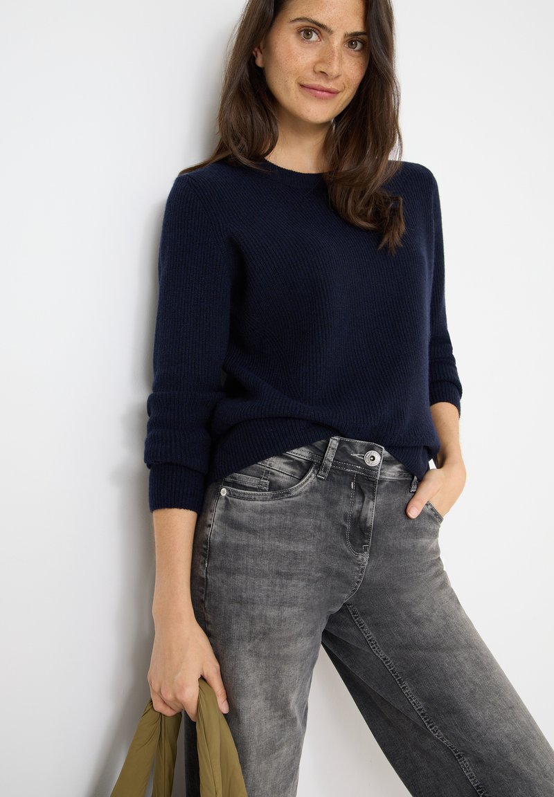 Marineblauwe ribgebreide trui met een ronde hals, gecombineerd met een paar grijze relaxed-fit jeans. Het model houdt een olijfgroene jas in een hand.