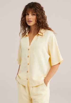 Femeie cu păr creț purtând o cămașă largă, de culoare galben pal, cu nasturi și pantaloni asortați, stând cu mâinile în buzunare, pe un fundal simplu.