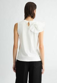 Top bianco senza maniche con scollatura rotonda, caratterizzato da un accento arricciato sulla spalla sinistra e una chiusura a bottone sul retro.