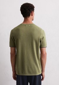 Koszula z krótkim rękawem w oliwkowym kolorze, o fakturowanym wykończeniu, z okrągłym dekoltem i kontrastowymi ciemnymi wykończeniami rękawów. Prosty, czysty design.