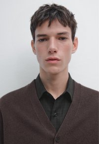 Brauner Cardigan über einem schwarz karierten Hemd, glatte Textur, V-Ausschnitt-Design, Knöpfe auf dem Hemd sichtbar, einfarbiger Hintergrund.