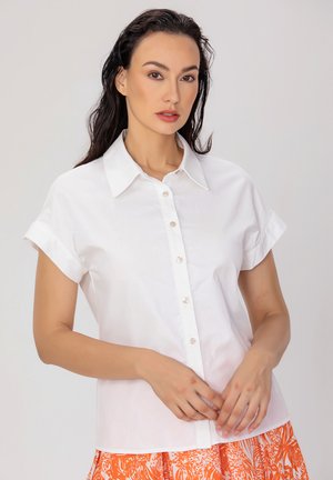 Chemise blanche à manches courtes avec un col, présentant une texture lisse et des boutons transparents, associée à une jupe orange à motifs.
