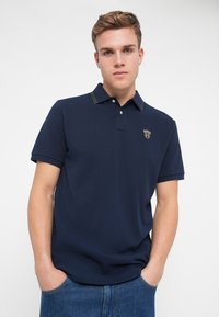 Sötétkék, rövid ujjú pólóing, texturált anyagból, két gombos nyakú, zöld díszvarrással. Kis logó található a mellkason.