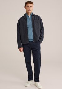 Tumesinine tõmblukuga jack, mille all on teal värvi kampsun, millel on krae ja tasku; koos tumedate pükste ja valgete tossudega. Sile tekstuur, juhuslik disain.