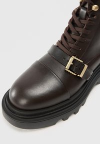 Bruine leren enkellaars met een ronde neus, veters en een decoratieve riem met een gouden gesp. Dikke zwarte zool met profielgroeven.
