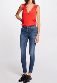 Top rojo sin mangas con detalles de encaje, combinado con jeans ajustados azules, cierre frontal con botones y zapatos negros con cordones sobre un fondo neutro.