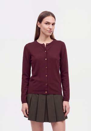 Un mannequin porte un cardigan bordeaux boutonné sur le devant et une jupe plissée foncée sur un fond blanc uni.