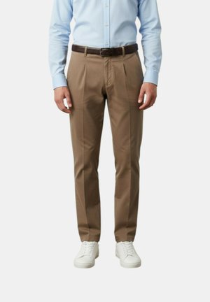 Uomo che indossa una camicia azzurro chiaro, pantaloni marroni plissettati con cintura scura e scarpe da ginnastica bianche, in piedi davanti a uno sfondo semplice.