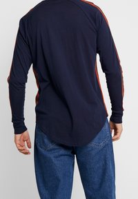 Marinblå långärmad skjorta med röda sidostråk, med rundad nederkant och mjukt bomullsmaterial. Avslappnad passform med en ledig design.