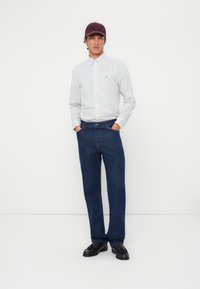 Camisa de botones a rayas en blanco y azul, jeans de denim oscuro y mocasines negros. Una gorra burdeos acentúa el atuendo. Hombre de pie contra un fondo blanco.