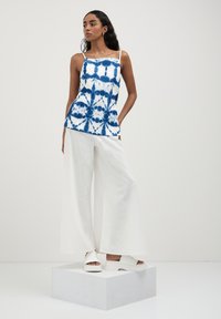 Top camicia bianco con motivo tie-dye blu, adornato con spalline sottili. Abbinato a pantaloni larghi bianchi e sandali bianchi.