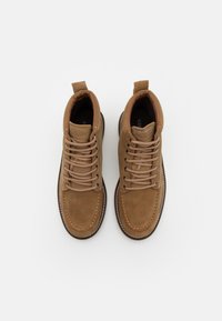 Botas de tobillo de ante marrón con cordones beige y costuras blancas en contraste. Cuentan con un bucle trasero para facilitar su uso y una suela acolchada.