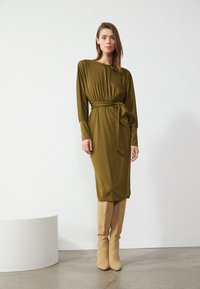 Olivengrønn, langermet kjole med smal midje og belte. Glatt stoff, kne-lengde, kombinert med beige, knelengde, semsket støvler.