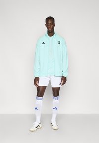 Mündiroheline kerge jope musta logoga, kombineeritud helesinise t-särgi, valgete pükste ja valgete sokkidega siniste triipudega.