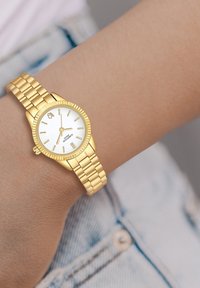 Goldene Armbanduhr mit einem runden weißen Ziffernblatt, goldenen Stundenmarkierungen und einer strukturierten Lünette. Verfügt über ein gliedertes Metallarmband.