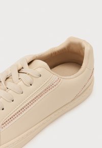 Heled helebeige tossud, mis on valmistatud siledast sünteetilisest materjalist, ümarate varvastega, paeltega esiosa, pehmendatud kraega ja dekoratiivse riba roosa kuldsete aktsentidega.
