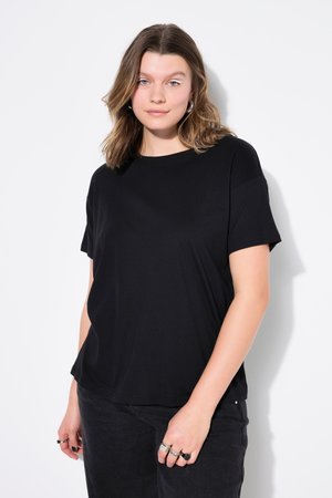 Młoda kobieta o brązowych włosach do ramion, ubrana w zwykłą czarną koszulkę i czarne dżinsy, stojąca na białym tle.