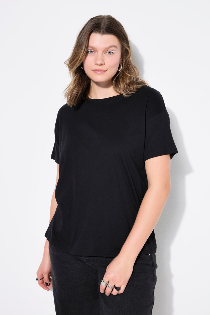 Joven con el pelo castaño hasta los hombros, vestida con una camiseta negra lisa y vaqueros negros, de pie frente a un fondo blanco.