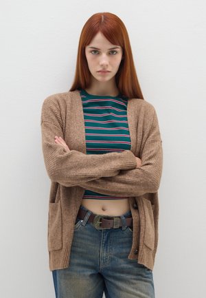 Cardigan marrone a coste con apertura frontale e tasche, indossato su una camicia a righe verdi, abbinato a jeans blu a vita alta e una cintura marrone.