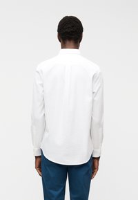 Άνδρας με λευκό πουκάμισο με μακριά μανίκια και μπλε παντελόνι, στέκεται με την πλάτη προς τα εμπρός σε απλό ανοιχτό φόντο.