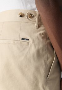 Gros plan d’un pantalon beige avec deux détails de boutons et une petite étiquette noire indiquant « BOSS » près de la poche arrière, porté avec une chemise blanche.