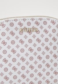 Bolsa texturizada blanca con el logo dorado "GUESS", que presenta un patrón repetido de logos y diseños florales en marrón claro. Detalle de cierre de cremallera.