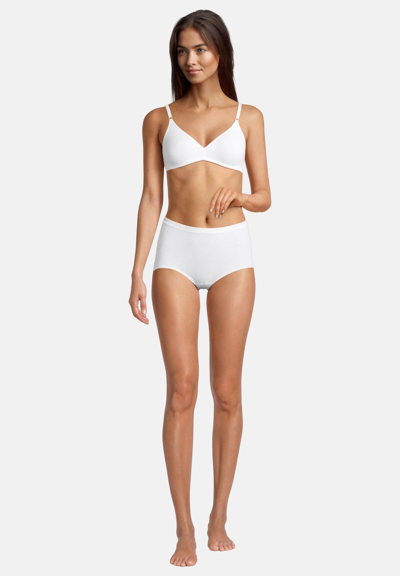 Junge Frau in einem weißen kabellosen BH und hochgeschnittenen Baumwollslips, die barfuß vor einem schlichten hellen Hintergrund steht.