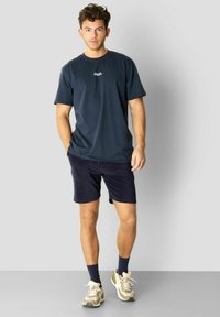 Marine t-skjorte med korte ermer og brystlogo, kombinert med marineblå shorts. Modellen har på seg beige sneakers og mørke sokker. Enkelt, uformelt design.